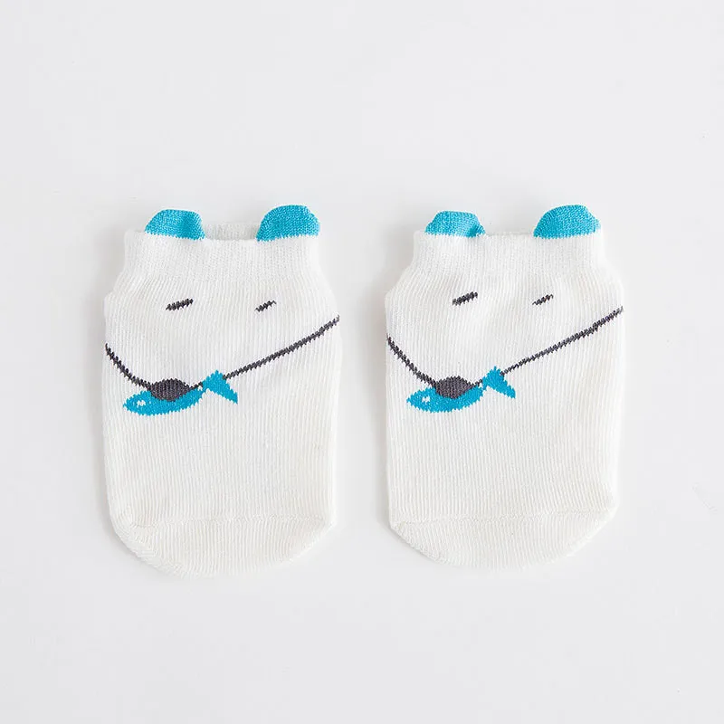 Un par de calcetines blancos para bebé con un bonito diseño. Cada calcetín tiene un motivo de pez azul con un pequeño detalle negro, y los calcetines están adornados con orejas azules en la parte superior, dándoles una apariencia divertida y animal.