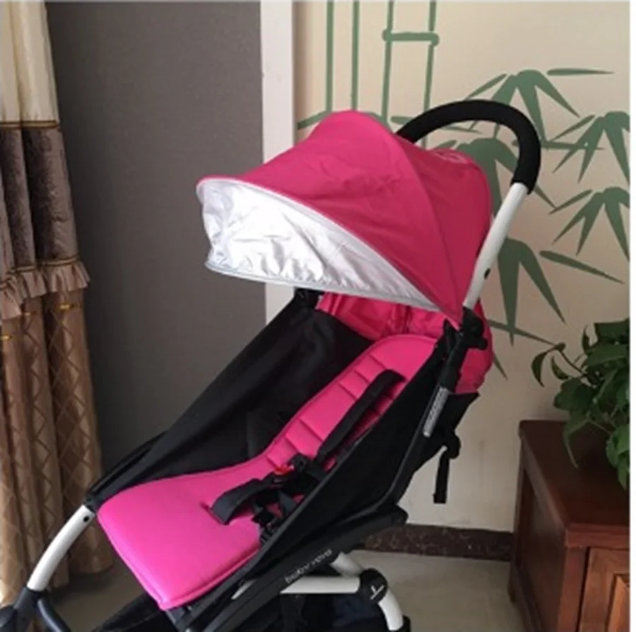 Un cochecito rosa con una capota parcialmente abierta. El cochecito tiene una estructura negra y parece estar diseñado para un niño pequeño, dado su tamaño y estilo. Está ubicado en el interior, con una pared con papel tapiz o decoración similar al bambú al fondo.