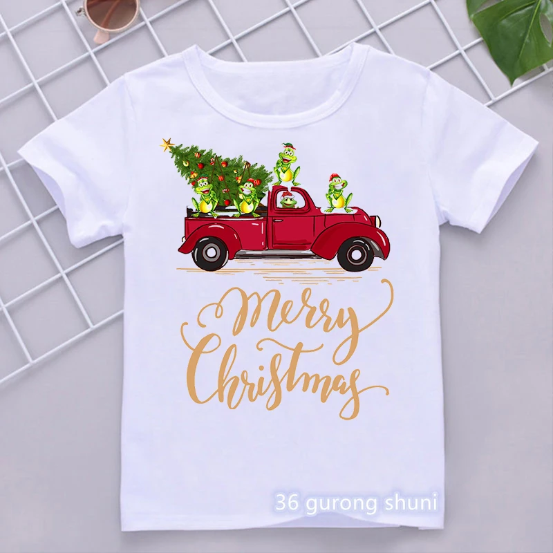 Una camiseta blanca con un diseño gráfico festivo. El diseño presenta un camión vintage rojo que lleva un árbol de Navidad con adornos y una estrella en la parte superior. Hay tres personajes de dibujos animados viajando en la parte trasera del camión, que parecen ser ranas con gorros de Papá Noel.