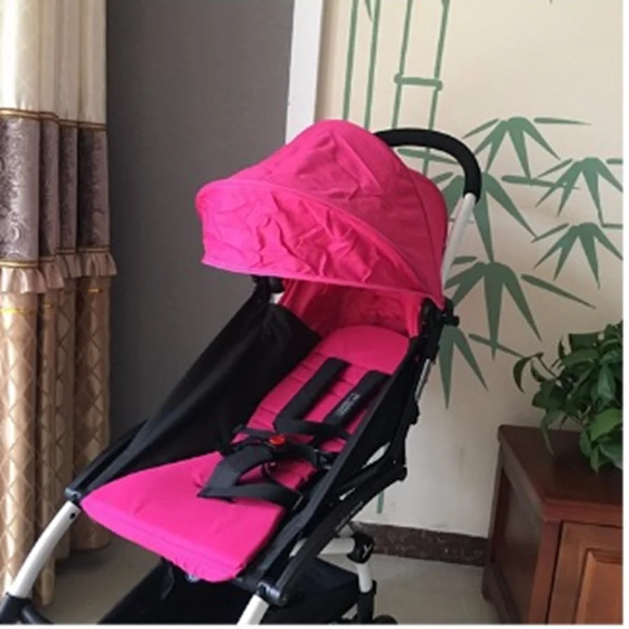 Un cochecito rosa con dosel. Parece ser un cochecito liviano y plegable, probablemente diseñado para uso urbano o para viajes. El cochecito está de pie y apoyado contra una pared con un papel pintado decorativo similar al bambú.