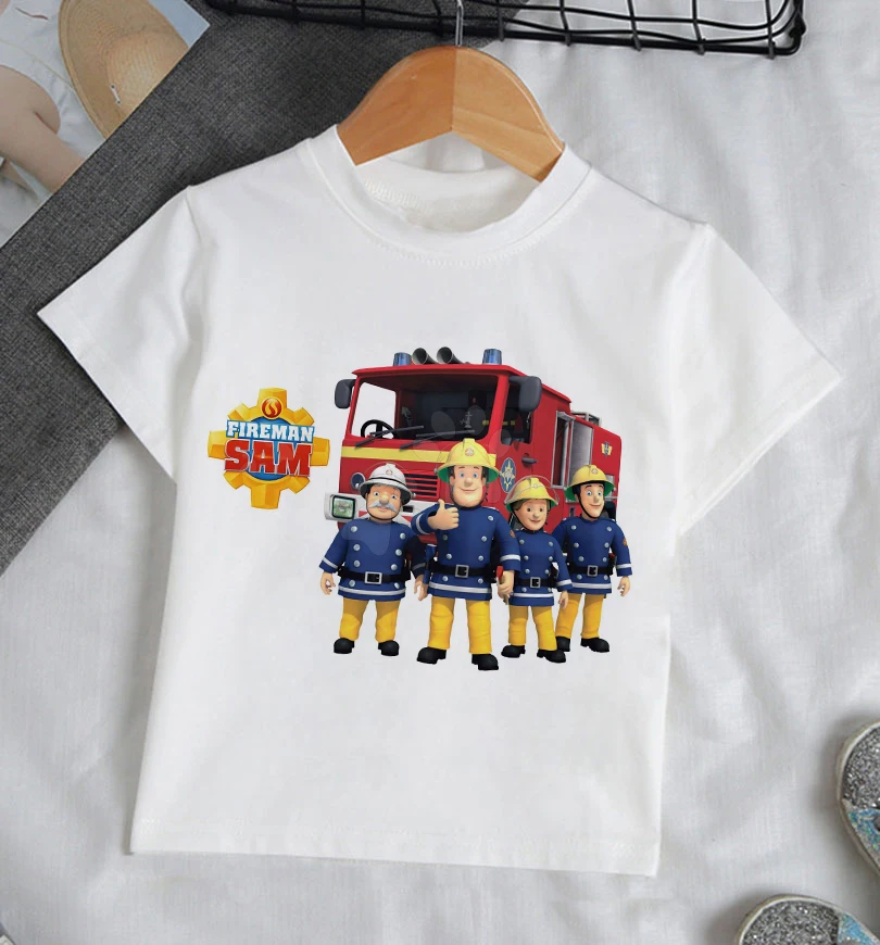 Una camiseta blanca con un diseño gráfico. El diseño presenta una caricatura de un camión de bomberos con las palabras FIREMAN SAM encima. Debajo del camión de bomberos, hay cuatro personajes de dibujos animados vestidos con uniformes de bombero, cada uno con una pose y expresión diferente.