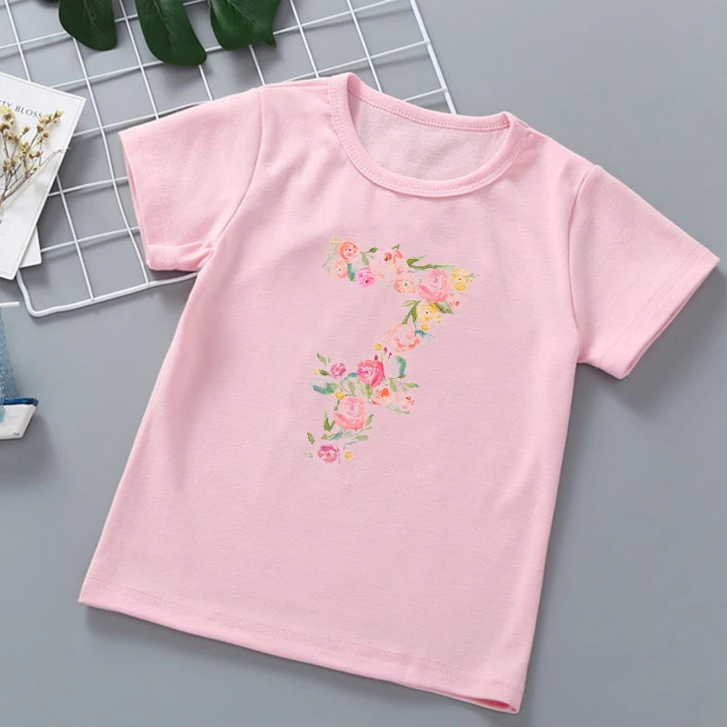 Camiseta rosa con diseño floral en la parte delantera. El diseño incluye un número 2 con flores y hojas alrededor, lo que sugiere que podría ser una camiseta de cumpleaños o relacionada con la edad, posiblemente para un niño que cumpla dos años.