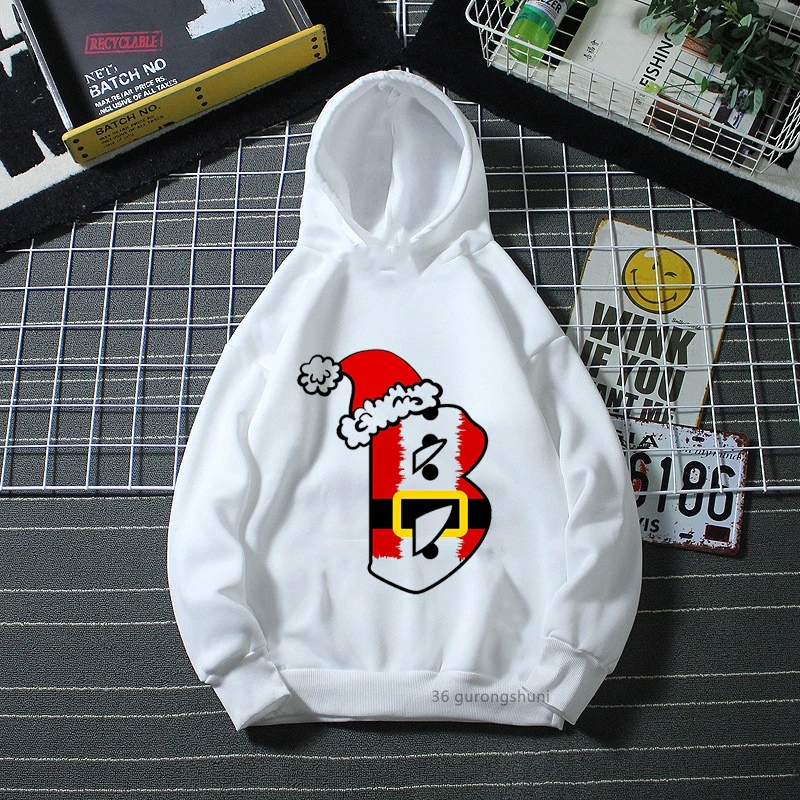 Una sudadera con capucha blanca con un diseño gráfico en el frente. El diseño presenta un gorro de Papá Noel rojo y blanco con un ribete blanco esponjoso y un pompón en la parte superior. Debajo del sombrero, hay un emblema en forma de escudo con un borde de rayas rojas y amarillas.