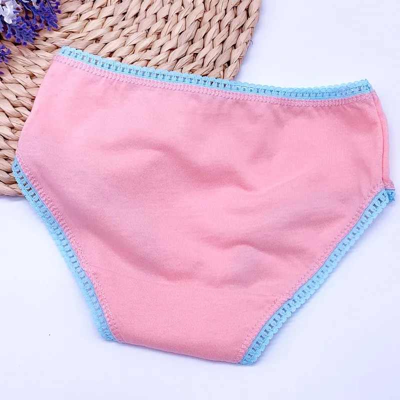 Un artículo de tela rosa con un borde azul, que parece ser una mascarilla. Está dispuesto de forma plana y es el foco principal de la imagen. Al fondo, hay una cesta o sombrero tejido y unas flores de color violeta, que dan un toque decorativo a la composición.