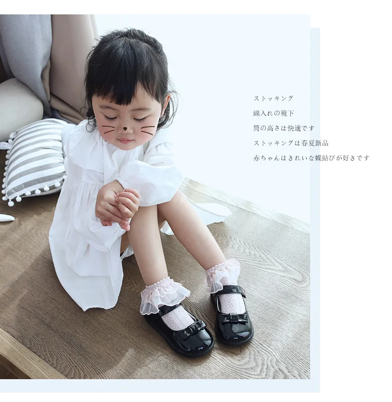 Un niño pequeño sentado en un suelo de madera. La niña lleva un vestido blanco con cuello con volantes y un par de zapatos negros con tiras blancas.