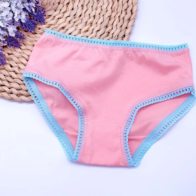 Un par de ropa interior de mujer de color rosa claro o coral y un ribete azul alrededor de la cintura y las aberturas de las piernas. La ropa interior parece estar colocada plana y hay una cesta tejida o un sombrero al fondo, junto con algunos elementos decorativos que pueden ser flores o bolas decorativas.