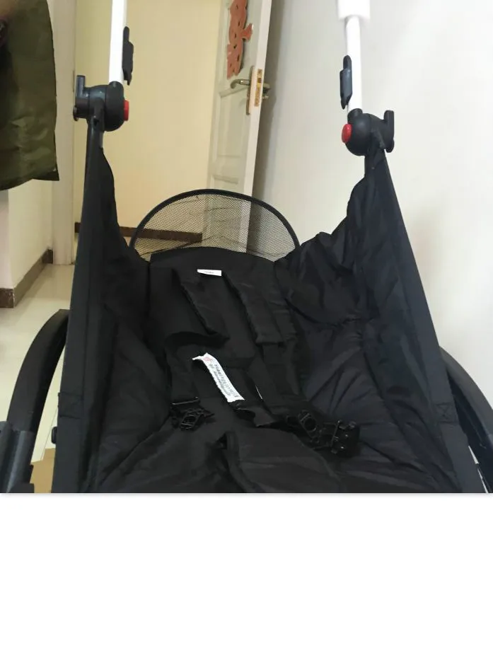 Un cochecito con la capota parcialmente abierta. El cochecito parece estar diseñado para bebés o niños pequeños, como lo indica la presencia de un sistema de arnés y la estructura general. Es probable que el dosel se utilice para proporcionar sombra y protección de los elementos al niño sentado en el cochecito.