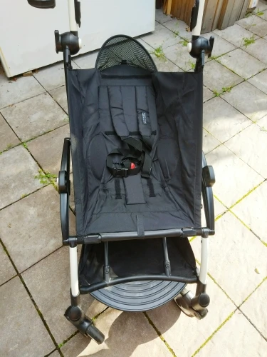 Silla plegable con asiento y respaldo de tela negra. La silla actualmente se encuentra plegada y apoyada contra lo que parece ser una pared o una estructura. La silla tiene un panel lateral de malla y una bolsa de tela negra fijada al respaldo.