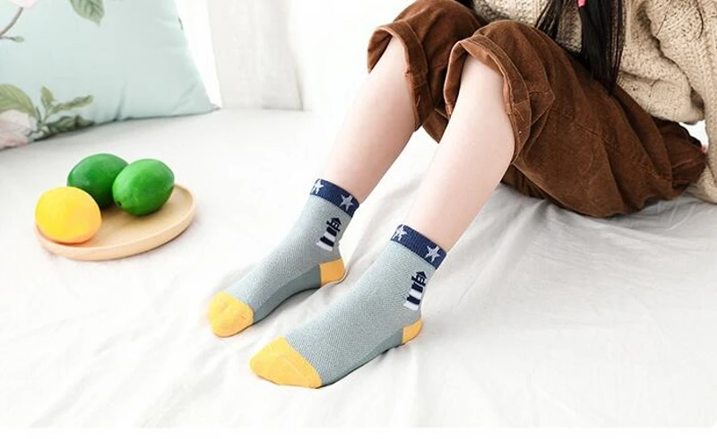 Una persona sentada en una cama con las piernas cruzadas. La persona lleva un par de calcetines grises con detalles en amarillo y azul, y pantalones marrones remangados hasta las rodillas.
