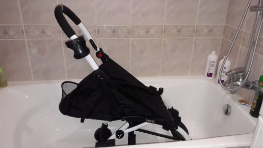 Un cochecito colocado dentro de una bañera. El cochecito parece ser compacto y plegable, y actualmente se encuentra plegado. La bañera está en un ambiente de baño, como lo indican las paredes de azulejos y el cabezal de ducha visible al fondo.