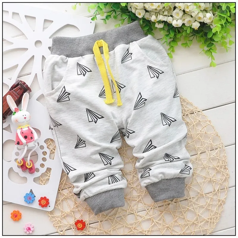 Pantalón infantil con fondo gris y estampado de triángulos blancos y negros. Los pantalones tienen un cordón amarillo en la cintura. Al fondo, hay algunos elementos decorativos como un tapete blanco, un pequeño conejito de juguete y unas flores artificiales.