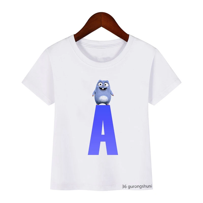 Una camiseta blanca con un diseño gráfico. El diseño presenta un personaje de dibujos animados que parece ser una criatura azul con una gran sonrisa y un cuerpo pequeño, sentado encima de una gran letra A azul.