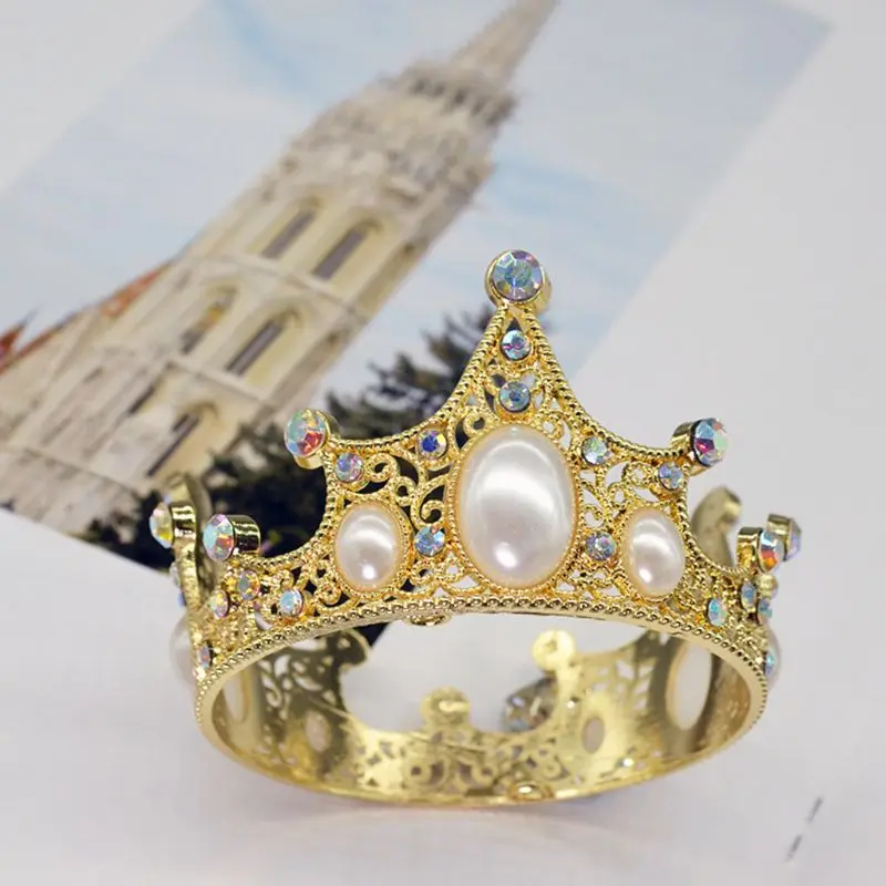 Una tiara dorada con un diseño que incluye perlas y lo que parecen ser piedras preciosas o cristales. La tiara está colocada sobre una superficie blanca, y al fondo hay una fotografía de un edificio con una torre alta, que podría ser una iglesia o un edificio histórico.