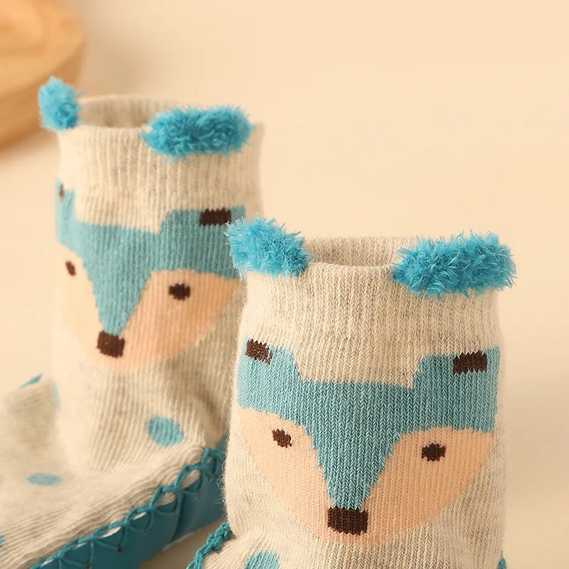 Un par de calcetines infantiles diseñados con apariencia de zorros. Tienen un color base blanco con detalles en azul y marrón para imitar el pelaje y los rasgos faciales de un zorro. Los calcetines también tienen pompones azules en la parte superior, que añaden un diseño divertido y lindo.