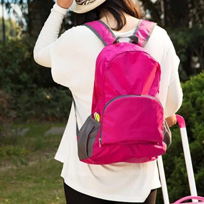 Una persona de espaldas, vestida con una mochila rosa y una camiseta blanca de manga larga. La persona también lleva un sombrero con un patrón. Parecen estar al aire libre, ya que hay árboles y hierba al fondo.