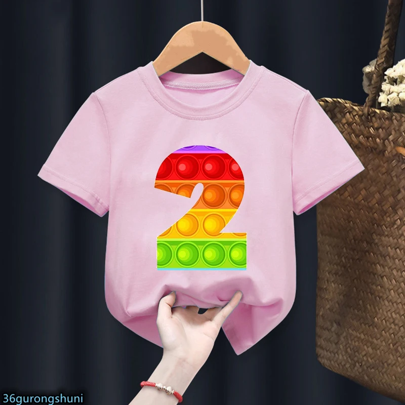 Una persona sosteniendo una camiseta rosa con un gráfico colorido. El gráfico parece ser un número 2 estilizado formado por círculos de colores, que posiblemente represente un diseño similar a Lego.