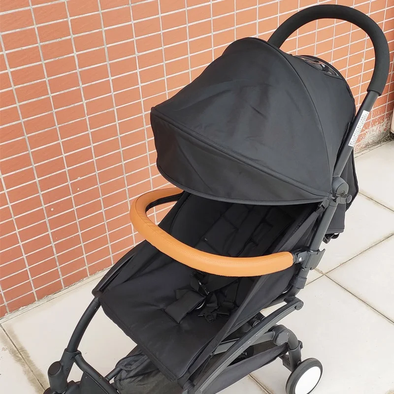 Un cochecito negro con dosel. El cochecito tiene una estructura y un asiento negros y parece estar diseñado para un solo niño. El dosel está levantado, brindando sombra al niño, y hay un manillar naranja visible que probablemente sirve como mecanismo de dirección.