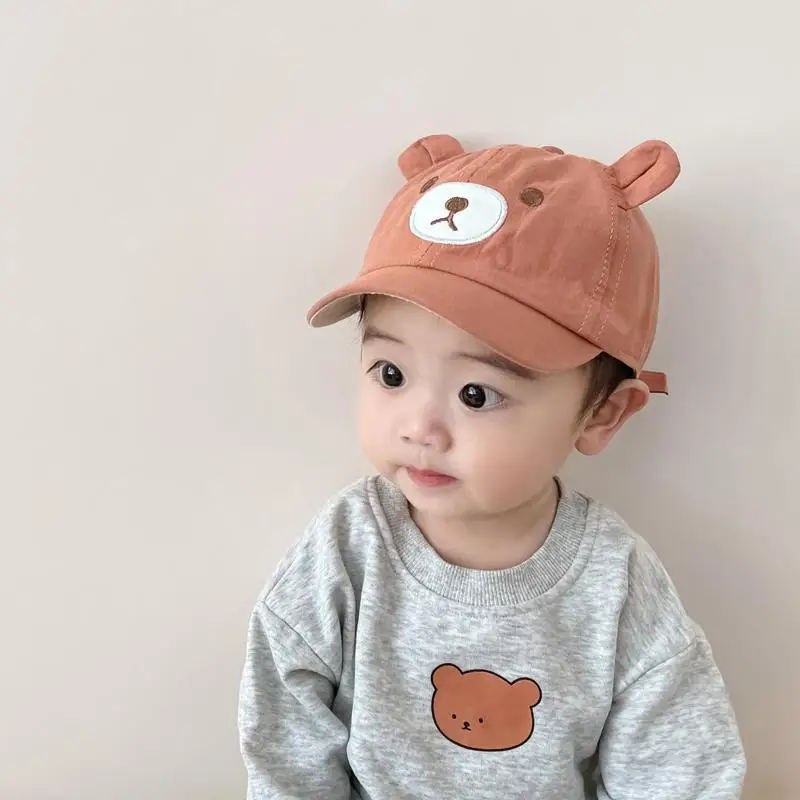 Un bebé con un sombrero marrón y una sudadera gris con un oso de peluche