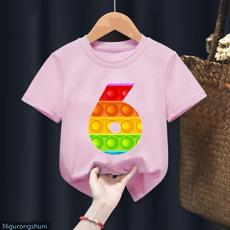 Una persona sosteniendo una camiseta rosa con un gráfico colorido. El gráfico parece ser un número 6 estilizado formado por coloridos ladrillos tipo Lego.