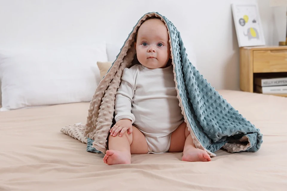 Un bebé sentado en una cama, parcialmente cubierto por una manta o un trozo de tela. El bebé viste un traje blanco y parece estar mirando directamente a la cámara con una expresión curiosa o contemplativa.