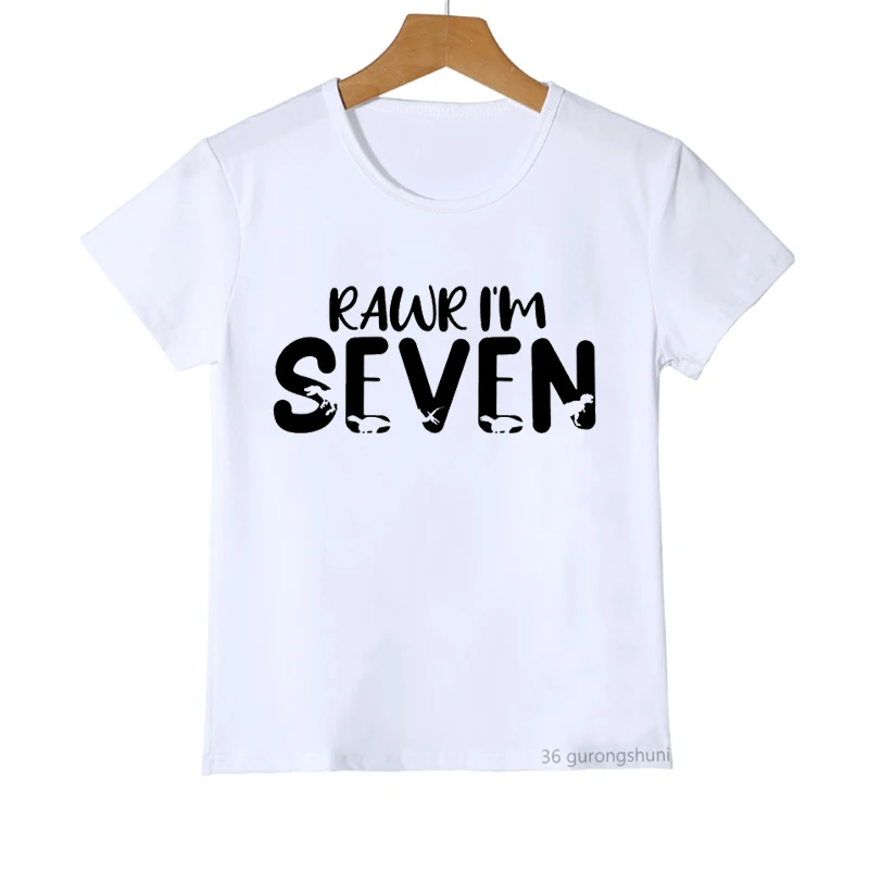 Una camiseta blanca con un diseño gráfico. El diseño presenta la frase RAWR IM SEVEN en una fuente divertida de estilo escrito a mano. Las letras son de color negro con un ligero efecto de sombra que les da una apariencia tridimensional.