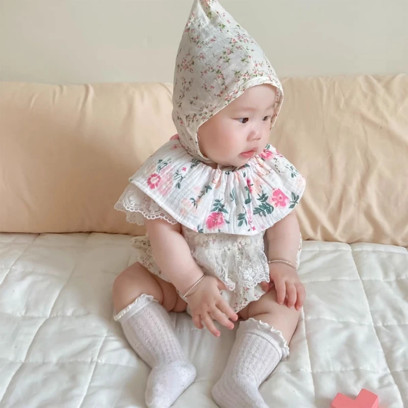 Un bebé sentado en una cama. La bebé lleva un vestido blanco con estampado de flores, una capota a juego y calcetines blancos. El bebé parece mirar hacia un lado con expresión tranquila.