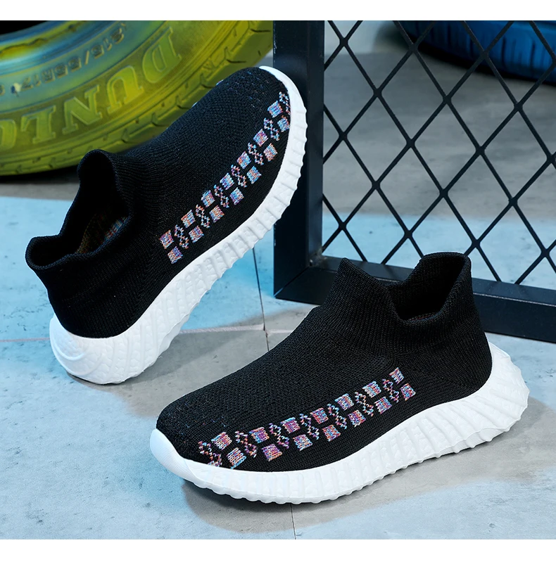 Un par de zapatillas negras con suela blanca. Las zapatillas tienen un diseño estampado en el lateral, que parece ser una serie de formas geométricas coloridas. El fondo incluye un objeto amarillo que podría ser un neumático, y hay una valla o puerta de metal negro.