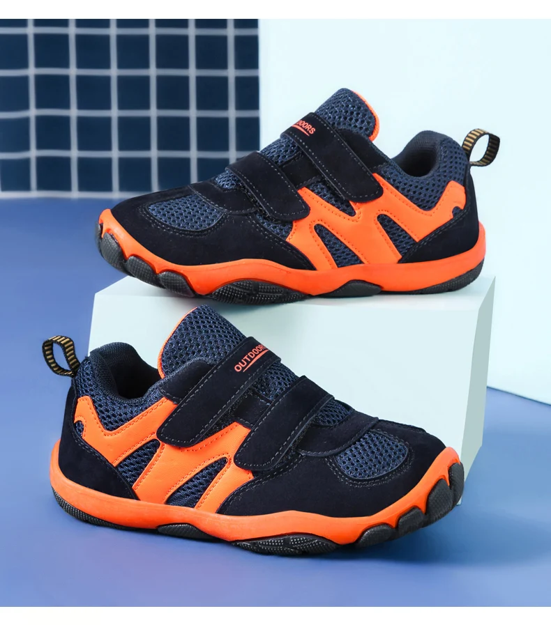 Un par de zapatos para niños. Son de color negro con detalles en naranja y parecen estar diseñados para actividades o deportes al aire libre, dada la suela resistente y el estilo general. La marca Outdoor es visible en el costado de los zapatos, lo que sugiere que están fabricados por una empresa con ese nombre.