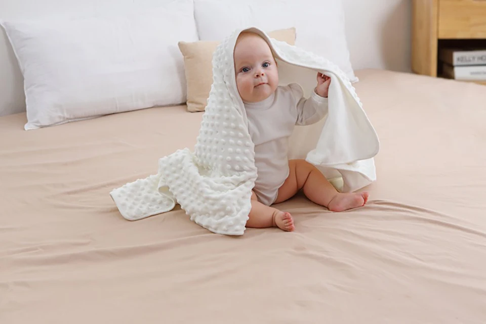 Un bebé sentado en una cama, sosteniendo una manta o sábana blanca sobre su cabeza. El bebé parece estar mirando directamente a la cámara con una expresión curiosa o juguetona. La cama tiene una colcha de color claro y al fondo hay almohadas.