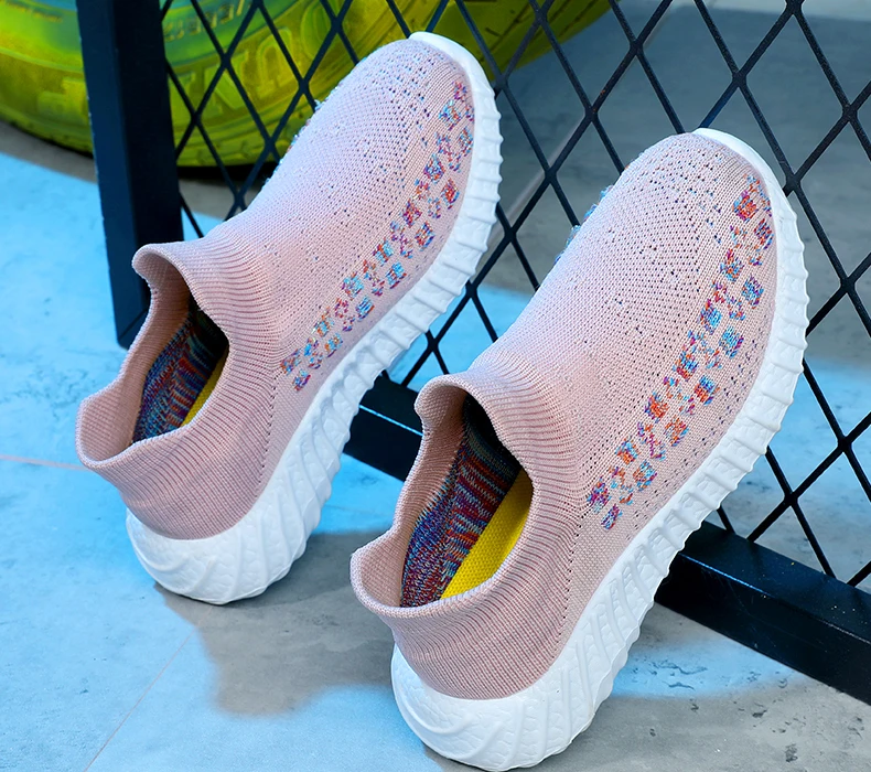 Un par de zapatillas con parte superior de punto. Las zapatillas tienen un color rosa pastel con un patrón de puntos o formas de colores en el lateral. Las suelas parecen ser blancas y gruesas, que es un diseño común para brindar comodidad y soporte.