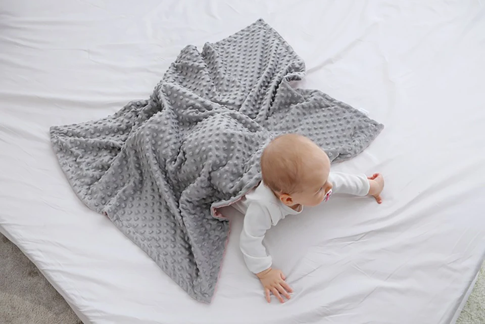 Un bebé acostado en una cama con una manta cubriéndolo parcialmente. El bebé parece llevar un traje blanco y mira hacia la cámara con una expresión curiosa. La manta tiene un estampado de pequeños puntos y la sábana es blanca, lo que contrasta con la manta.