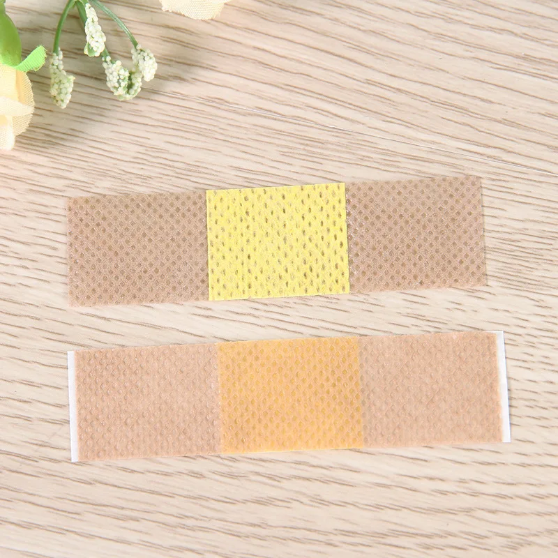 Dos vendas adhesivas colocadas sobre una superficie de madera. Cada vendaje tiene un centro amarillo con un patrón de pequeños agujeros, que probablemente esté diseñado para permitir que el aire circule y promover la curación.