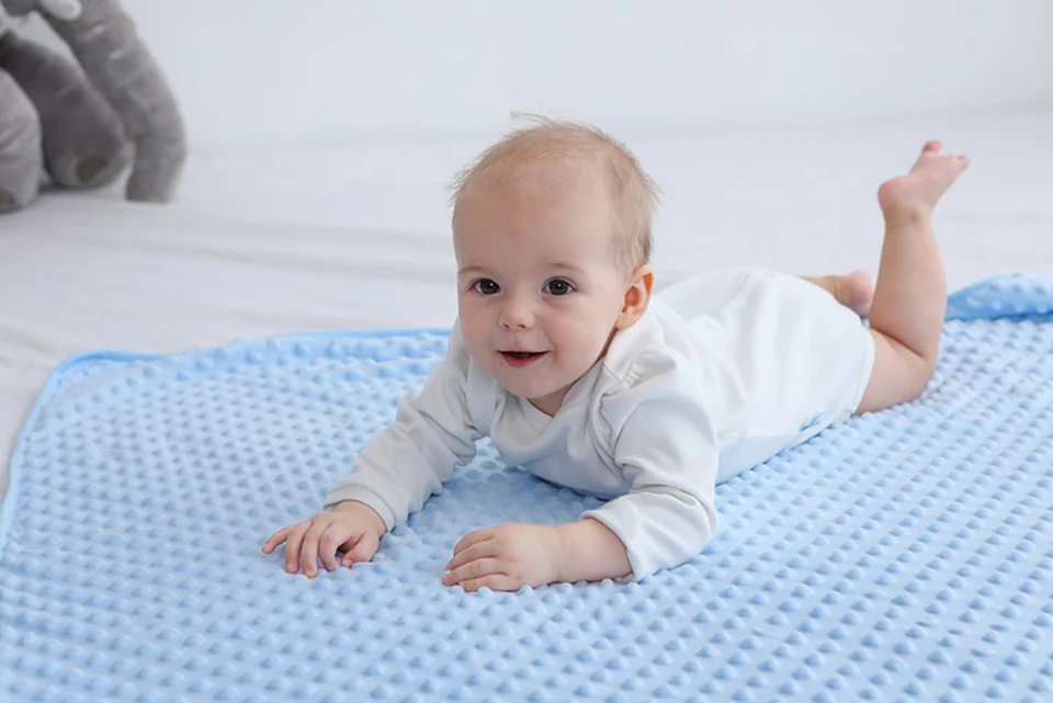 Un bebé acostado sobre una alfombra de juego azul. El bebé parece estar sonriendo y mirando directamente a la cámara. Al fondo, hay un animal de peluche que parece un elefante gris, lo que añade un elemento lúdico a la escena.